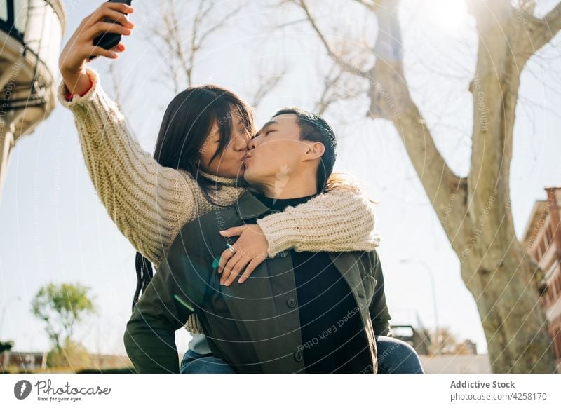 Loving young Asian couple taking selfie while cuddling and kissing in park cuddle love date affection smartphone together hug embrace romantic bonding