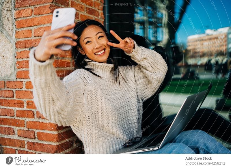 Positive Asian female millennial taking selfie while using laptop on street woman smile smartphone work freelance cheerful two fingers internet social media