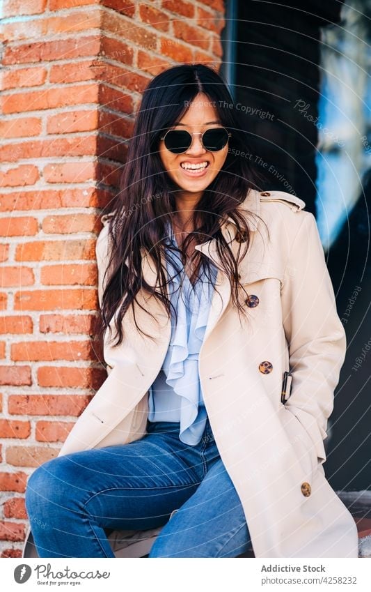 Trendy young Asian female sitting near brick building on city street woman trendy style confident personality cheerful charismatic sunglasses positive