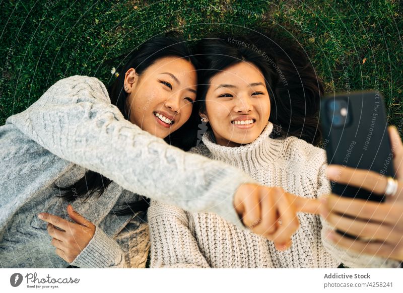 Joyful young ethnic female friends lying on lawn and showing tongues while taking selfie women show tongue joy happy smartphone cheerful having fun spend time