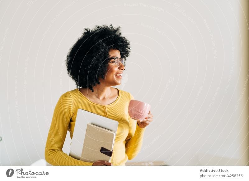 Black woman in eyeglasses with cup and notebook with laptop smart cheerful smile positive mug coffee hot drink female yellow sweater modern black relax work