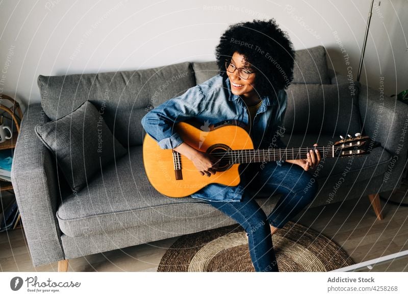 Positive creative black woman playing guitar on sofa in lounge positive smile cheerful acoustic home music female eyeglasses casual instrument african american