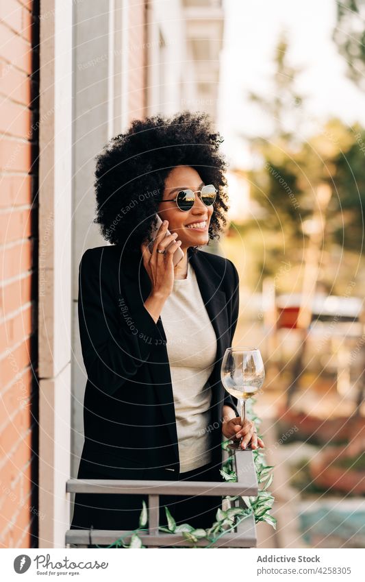 Stylish black woman in trendy sunglasses talking on smartphone style suit elegant phone call wine positive female smile cheerful mobile alcohol balcony lady