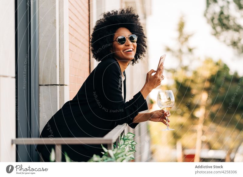 Stylish black woman in trendy sunglasses using smartphone style suit elegant wine positive female smile cheerful mobile alcohol balcony lady confident afro