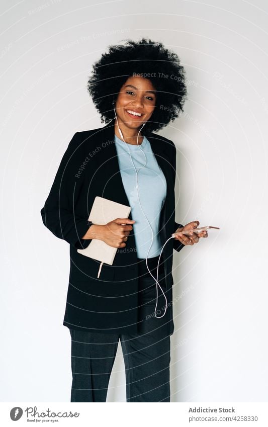 Cheerful black woman with notebook and smartphone listening to music cheerful smile happy earphones mobile positive female elegant suit formal outfit
