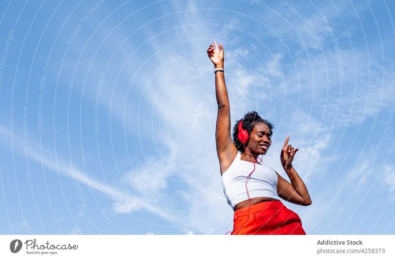 Excited black woman listening to music in headphones and dancing dance joy having fun blue sky melody enjoy arms raised happy young fit energy positive