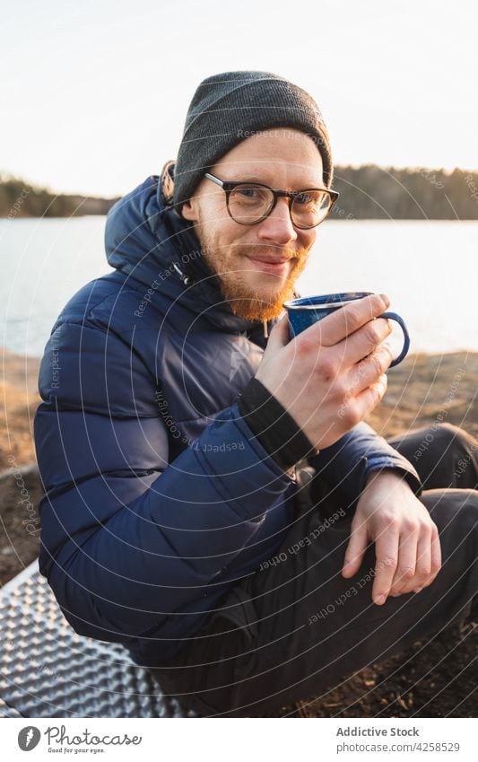 Happy young guy drinking hot beverage during camping on lake shore man coffee hiker travel rest positive smile male beard style warm clothes eyes closed autumn