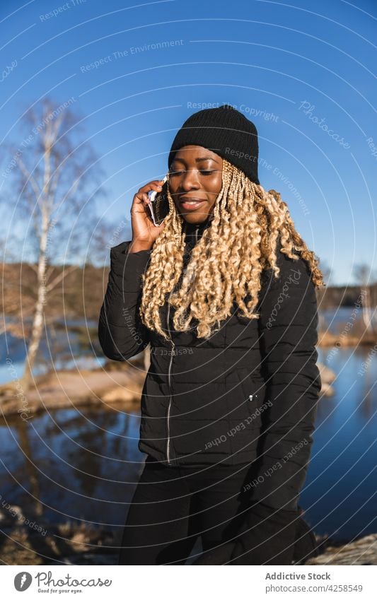 Black woman standing on shore near pond smartphone talk using communicate speak river coast nature listen female mobile gadget smile outerwear cellphone