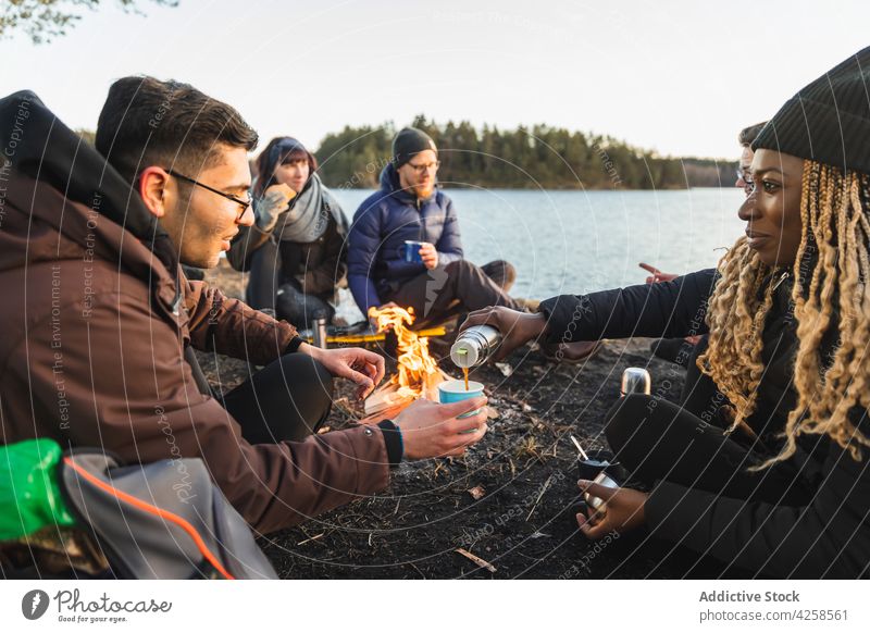 Black woman pouring tea from thermos sitting near bonfire women friend coast river cup male burn female drink together beverage hot friendship campfire