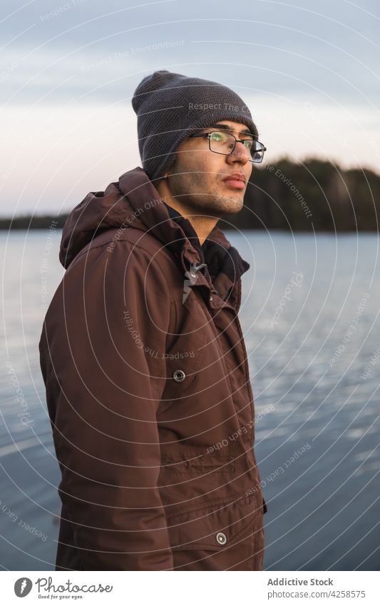 Young man in outerwear standing near rippling river dreamy coast peaceful nature environment freedom harmony silent male casual lake shore water serious beard