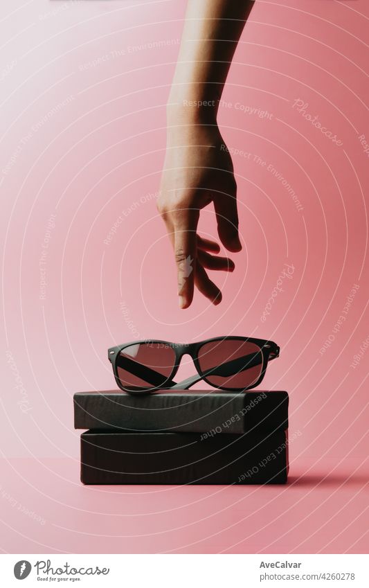 Hand reaching a pair of sunglasses over a pastel pink background with copy space minimal concept stylish frame plastic modern hipster minimalism female fun