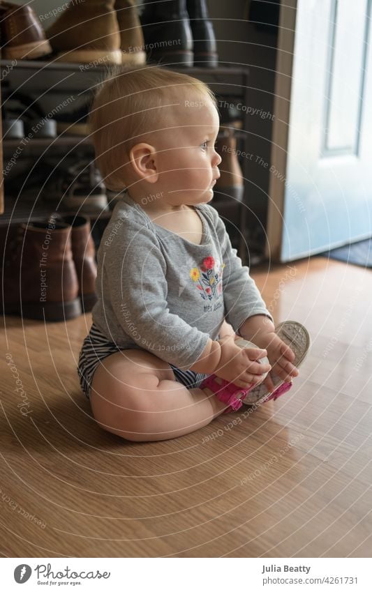 Toddler seated in front of a shoe rack in her home while trying on first pair of shoes; child sits upright with good posture holding feet together in butterfly pose