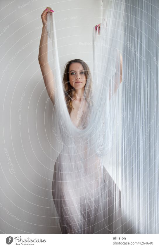 muscular beautiful young attractive sexy woman by the window, in a body flattering fashionable moving dress from the threads of a falling thread curtain white