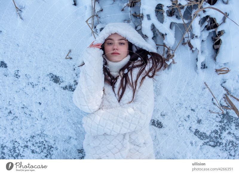 portrait of beautiful, young, secy, cute, brunette woman, freezing in the cold winter in white coat and hood, lying surreal like standing on the frozen lake, full ice flowers, copy space
