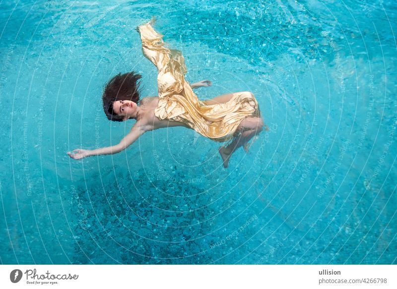 beautiful sexy young woman mermaid in gold dress, cloth floats weightless elegant in the blue turquoise spa pool water dreamy floating pensive unworldly
