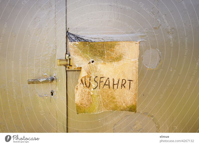 A square, torn piece of leather with the scrawled inscription " AUSFAHRT " covers the door latch on a closed, khaki-colored metal garage door whose rust stains have been painted over / makeshift