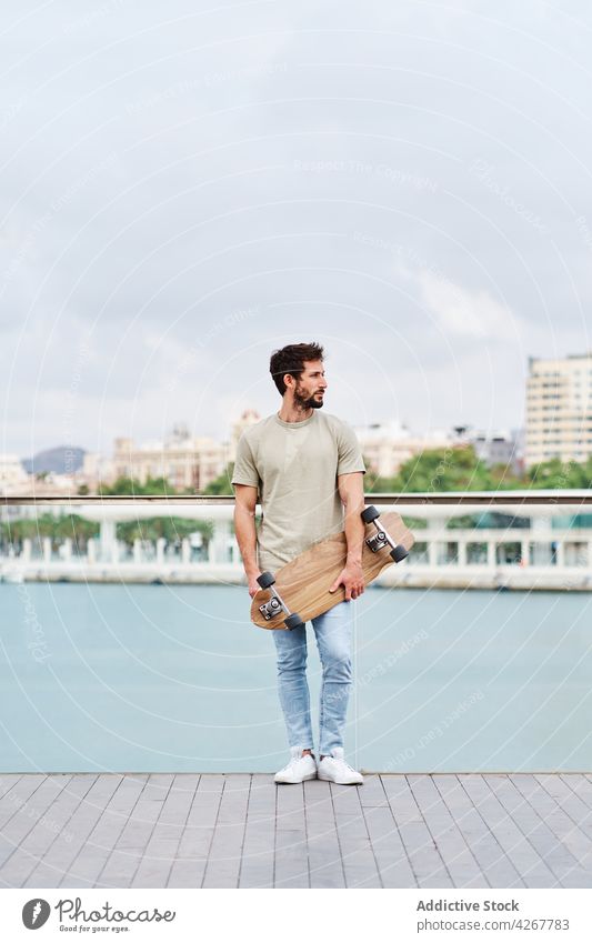 Bearded man with skateboard standing on city lakeside skater unemotional casual waterfront promenade embankment young river pond summer sunny town boardwalk