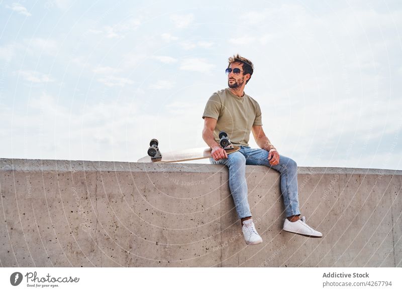 Cool man with skateboard resting on border on embankment sea chill shore waterfront masculine carefree relax male summer seafront leisure young free time coast
