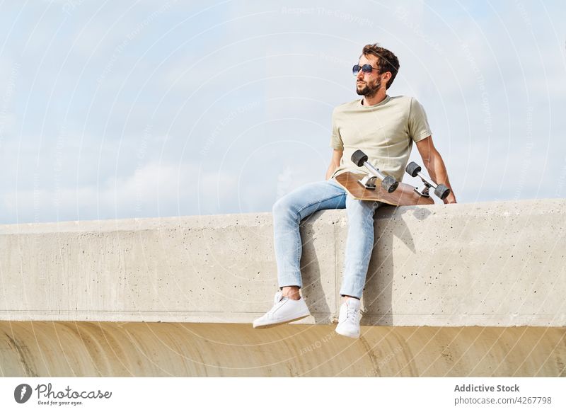 Cool man with skateboard resting on border on embankment sea chill shore waterfront masculine carefree relax male summer seafront leisure young free time coast