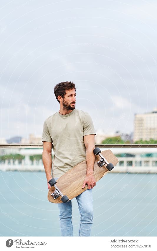 Bearded man with skateboard standing on city lakeside skater unemotional casual waterfront promenade embankment young river pond summer sunny town boardwalk