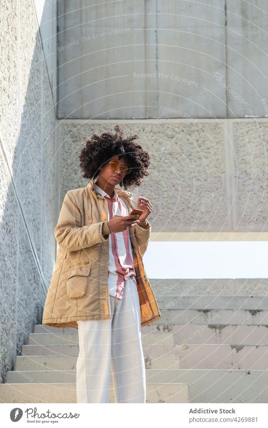 Black man in retro clothes chatting on smartphone on stairs text messaging hand in pocket vintage style afro internet using gadget device coat online watching
