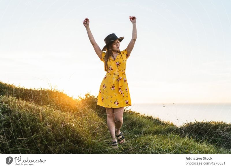 Young woman standing with arms raised on grassy lawn sundress countryside nature dreamy grassland meadow young field hat appearance serene grace feminine