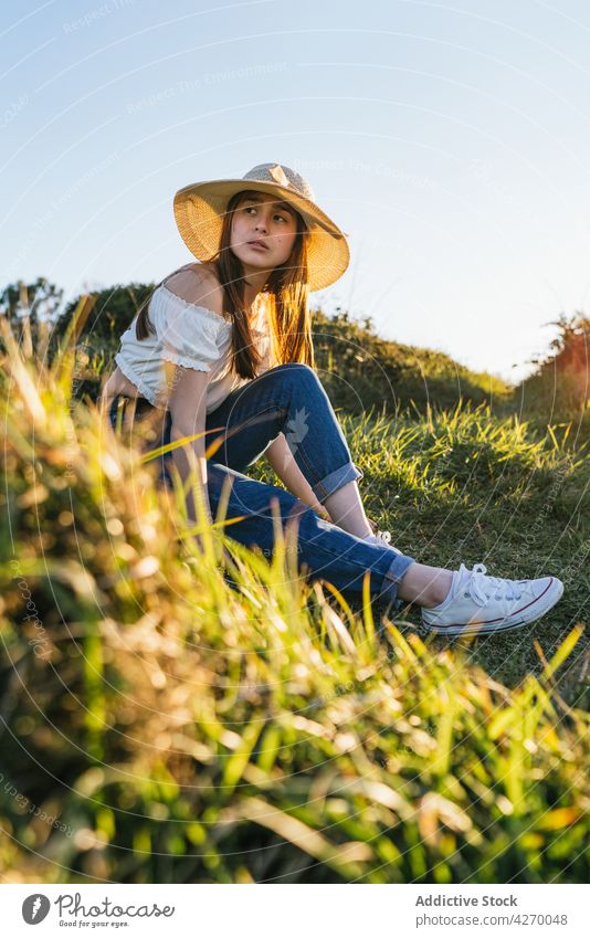Young woman siting on grassy meadow in sunny countryside lawn dreamy peaceful summer rest nature serene trendy appearance tranquil jeans field calm hat casual