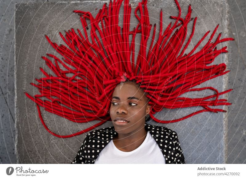 Black woman with bright hairstyle lying on concrete surface braid red cool carefree serious gaze female individuality personality vivid ground long hair