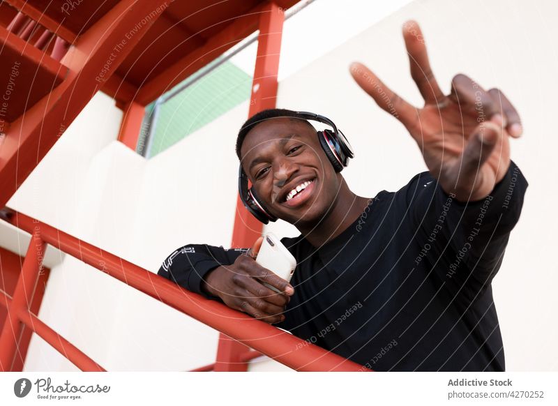 Cheerful black man in headphones showing V sign v sign toothy smile cheerful two fingers listen gesture carefree satisfied positive staircase demonstrate modern