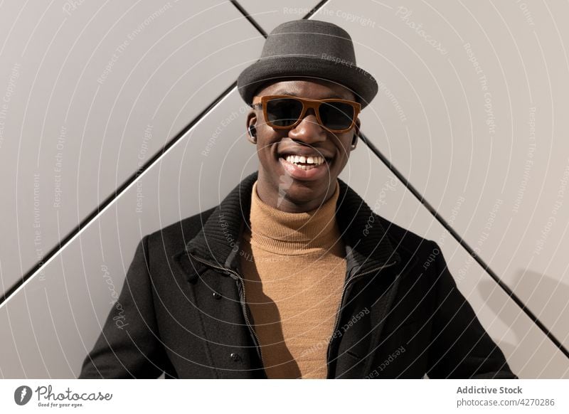 Stylish cheerful black man standing against white wall style outfit confident appearance trendy cool well dressed hat sunglasses individuality personality