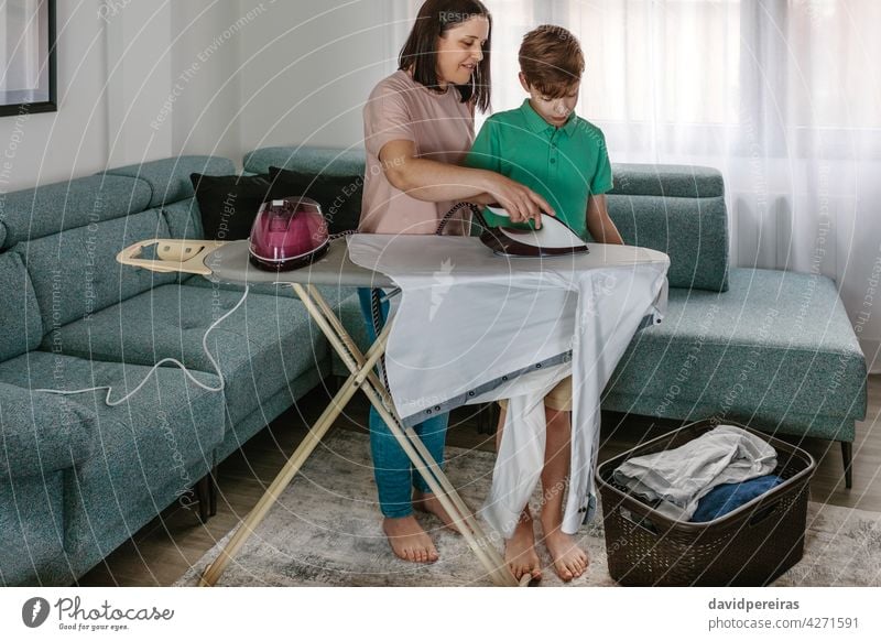 Mother teaching her teen son to iron mother ironing home helping ironing board laundry basket clothes together living room housewife teenager kid woman child