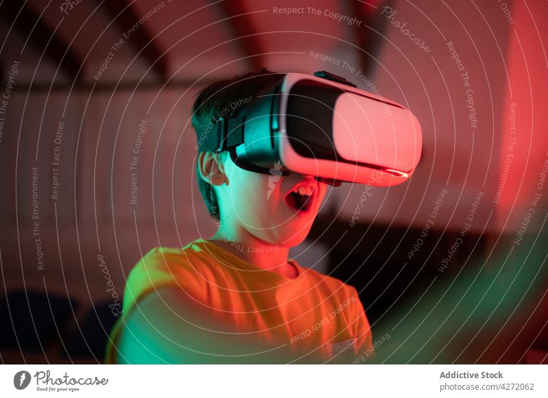 Cute child in VR goggles playing video games virtual reality entertain innovation vr boy digital equipment fun surprised kid casual neon illuminate darkness