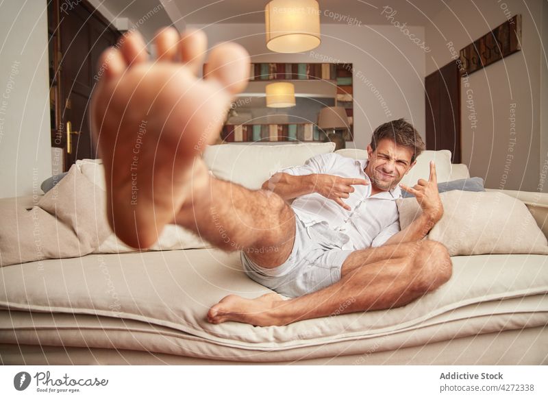 Joyful young guy winking and showing horn gesture while resting on sofa man sign of horn relax couch having fun positive free time joy happy comfort cozy male