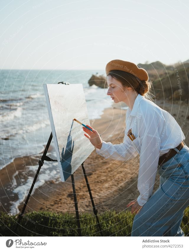 Female artist on shore near sand and sea while painting woman draw hobby canvas female young easel beret eyes closed creative artwork style white shirt jeans