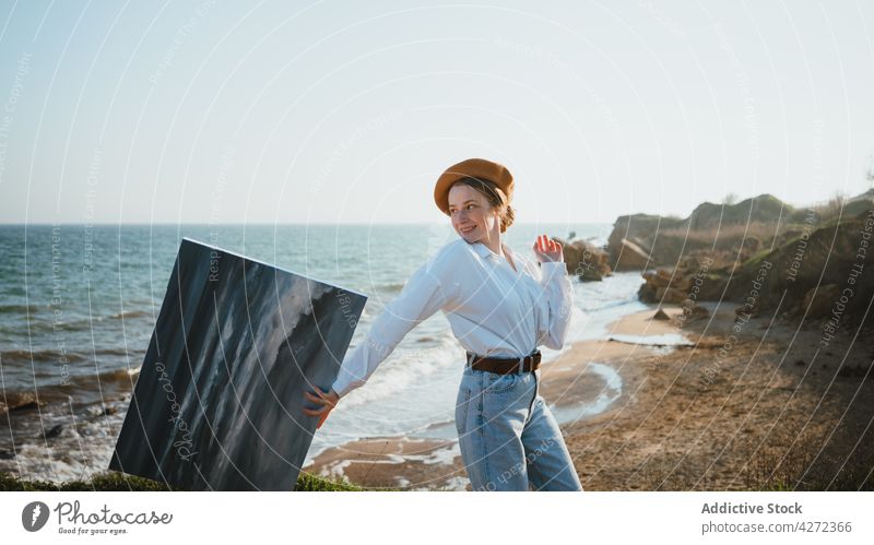 Smiling young woman walking on seashore with painting in hand nature content happy painter beach style positive coast female trendy hat enjoy ocean smile
