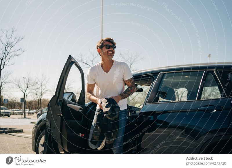 Smiling tattooed man getting out of automobile in sunlight driver get out cheerful masculine macho sunglasses style modern car brutal unshaven sky natural
