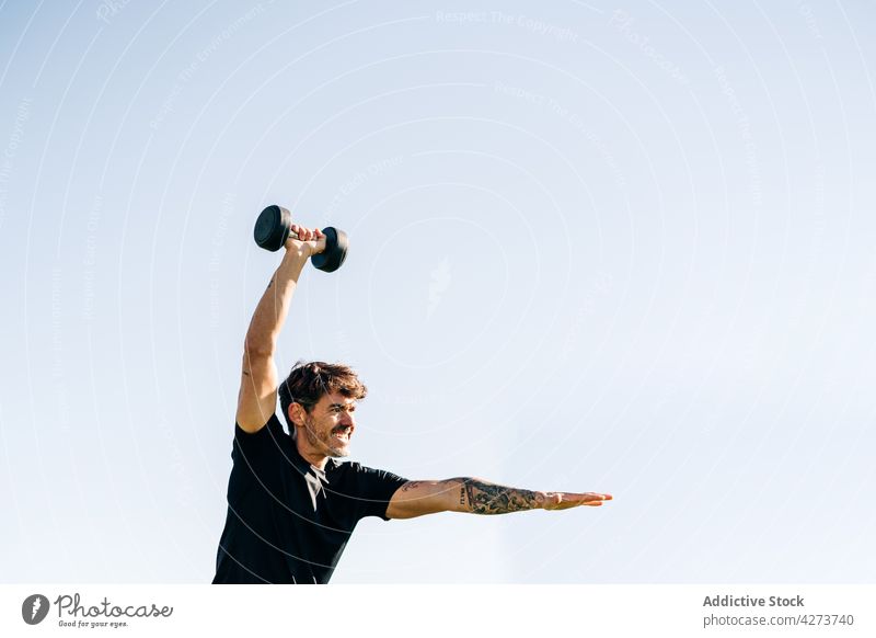 Tattooed sportsman exercising with dumbbell in sunlight arms raised workout training endurance motivation tattoo tension weight sky outstretch activity