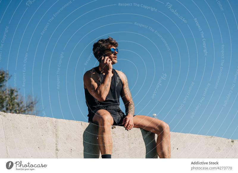Sportsman talking on smartphone on fence in town sportsman speak voice spare time break using gadget device cellphone shadow sit park shade communicate athlete