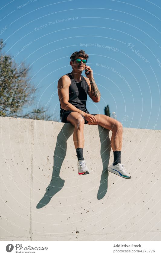 Sportsman talking on smartphone on fence in town sportsman speak voice spare time break using gadget device cellphone shadow sit park shade communicate athlete