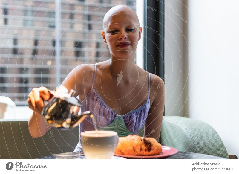 Hairless woman pouring tea from teapot in cup near pastry drink table positive cafe hairless female young window bald smile sofa room cafeteria food plate bun