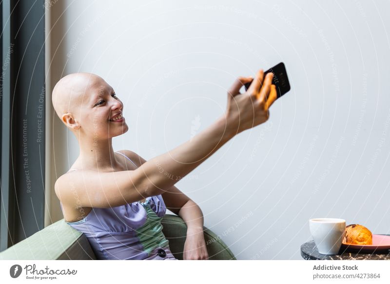 Bald woman taking selfie with smartphone on couch near table with cup sofa bald drink room modern female sit using alopecia gadget surfing device cellphone
