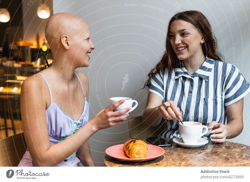 Cheerful young female friends having fun while eating croissant and drinking coffee in cafe women coffee break laugh joy positive enjoy cheerful humor alopecia
