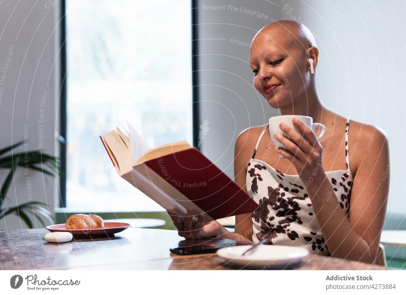 Positive young hairless woman drinking coffee and reading novel in cafeteria book alopecia relax smile enjoy education bookworm female bald disease literature