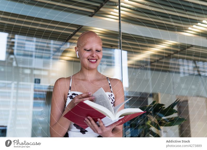 Smiling young hairless lady reading book in workplace woman smile alopecia happy positive story office style personality female bald trendy tws earphones listen