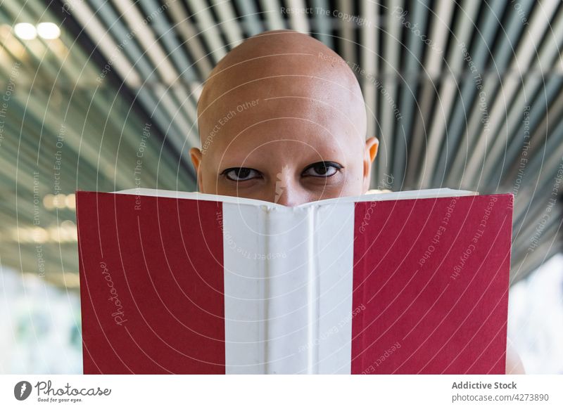 Young hairless woman hiding behind book and looking at camera read alopecia gaze interest hide disease novel education knowledge female young bald makeup
