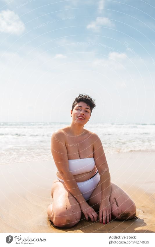 Lady sitting on sandy coast near sea woman beach ocean smile plus size swimsuit vacation blue sky happy female young aqua enjoy alone bright toothy smile