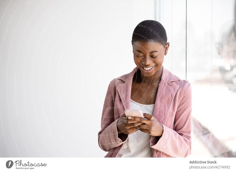 Cheerful black woman using smartphone against glass wall trendy fashion cheerful browsing contemplate message candid gadget device cellphone text reflection
