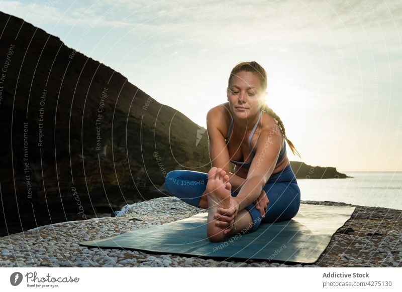 Woman performing forward bend on yoga mat against sea woman stretch hands clasped touch feet concentrate seashore sunshine pebble healthy lifestyle lean forward