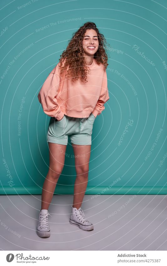 Cool youngster standing with hands in pockets in studio woman fun style cool posture glad carefree positive millennial appearance female curly hair cheerful