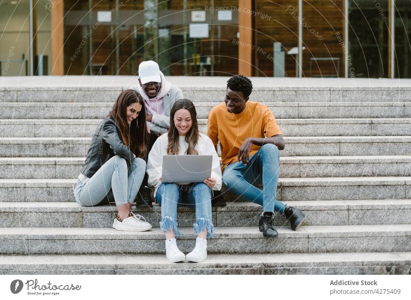 Multiethnic students using laptop on steps of university men women friend watch internet share glad enjoy male female together online cheerful friendship